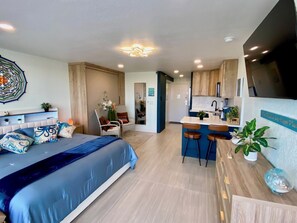 A modern hotel room with a large bed, a sitting area with a chair and a small table, a kitchenette, and a bathroom visible through an open door. - Stunning Brand New Studio - Balcony w/ Scenic Ocean and Sunset Views-Heated pool (Daytona Beach)