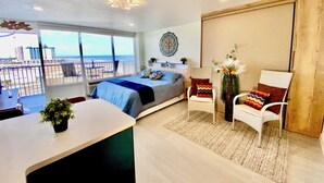 Une chambre moderne avec un grand lit, un balcon offrant une vue sur la plage et les bâtiments, un bureau agrémenté d’une plante et une chaise avec un coussin décoratif.