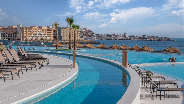 Ein Poolbereich mit Liegestühlen, einer geschwungenen Poolkante und Blick auf das Meer sowie Gebäude im Hintergrund.