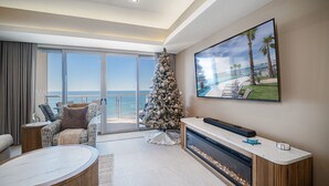 Ein modernes Wohnzimmer mit großem Fenster und Meerblick, einem Kamin, einem Weihnachtsbaum und einem großen Fernseher, der eine Strandansicht zeigt.