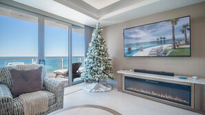 Ein modernes Wohnzimmer mit großem Fenster mit Meerblick, einem Kamin, einem Weihnachtsbaum und einem Fernseher, der einen Blick auf ein Resort zeigt.