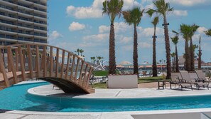 Un área de piscina con un puente de madera, sillones de descanso y palmeras.
