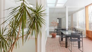 Uma sala de jantar moderna com uma mesa de vidro, cadeiras de metal, um vaso de planta e vista através de uma porta de vidro.