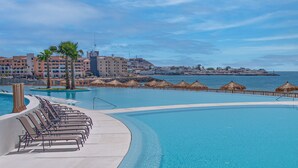 A resort pool with lounge chairs, palm trees, and a view of the sea and buildings. - One 1502 Hotel Room – New Seaside Gem Delivering Style, Comfort, and Panoramic Views (Puerto Peñasco)