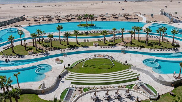 Un grand complexe de piscines à plusieurs niveaux, agrémenté de palmiers, de chaises longues et d’une plage en arrière-plan.