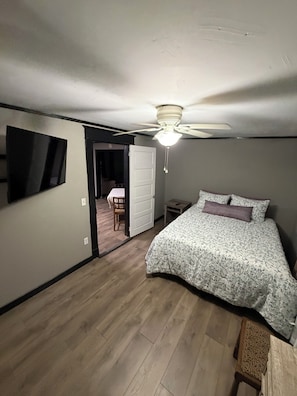 Une chambre à coucher avec un lit, un ventilateur de plafond, une télévision et une porte donnant sur une autre pièce.