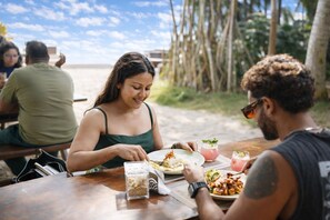 A beachside dining area with wooden tables and chairs, a clear sky, and a sandy beach in the background. - Noa Sands Talpe  by Ceylon Vistas (Talpe)
