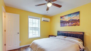 A bedroom with a bed, a ceiling fan, a window, an air conditioning unit, and a painting on the wall.