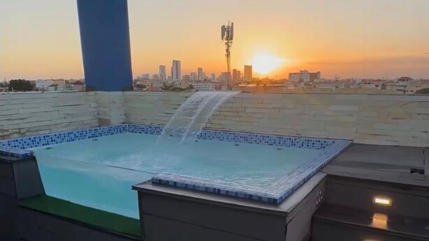 Ein Pool auf dem Dach mit Wasserfall, umgeben von einer Stadtlandschaft bei Sonnenuntergang.