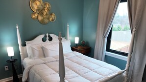 Een slaapkamer met een hemelbed, een nachtkastje met een lamp, een decoratief object aan de wand en een raam met gordijnen.
