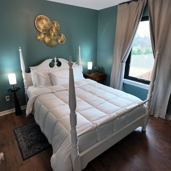 A bedroom with a four-poster bed, a nightstand with a lamp, a wall-mounted decorative item, and a window with curtains.