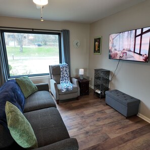 A living room with a grey sofa, a small armchair, a TV on the wall, and a window with a view of a yard.