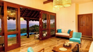 A spacious living area with a wooden floor, a sofa, a chair, a table, and a view of a pool and mountains through large glass doors. - Kandy Knightsdale Villa (Menikhinna)