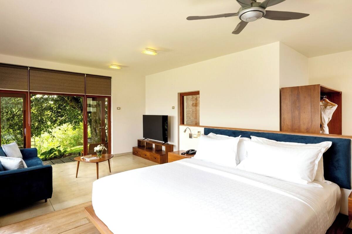 Une chambre à coucher moderne, dotée d’un grand lit, d’un ventilateur de plafond, d’une télévision et offrant une vue sur la verdure grâce à de grandes fenêtres.
