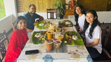 Un grupo de personas sentadas alrededor de una mesa con diversos alimentos y bebidas, bajo una estructura de techo de madera.