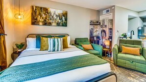Ein Hotelzimmer mit einem großen Bett, einem grünen Sessel, einer grünen Couch, einem Stadtbild als Gemälde und einer Küchenzeile.