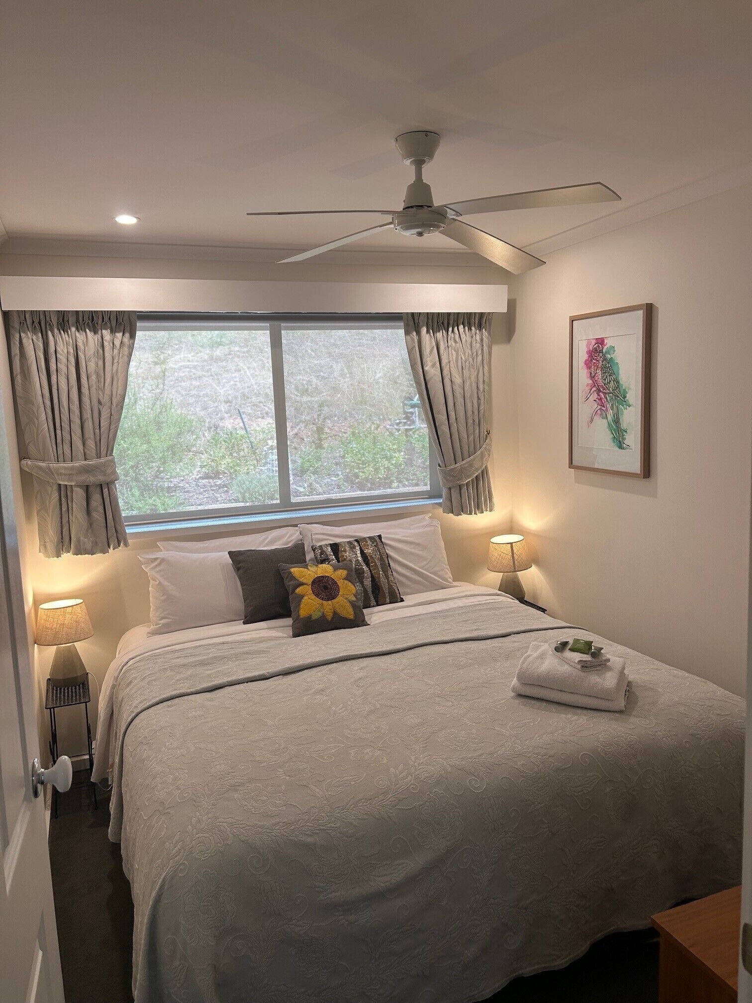 Une chambre à coucher comprenant un lit, un ventilateur de plafond, une fenêtre avec des rideaux, une lampe de chevet et une œuvre d’art encadrée accrochée au mur.