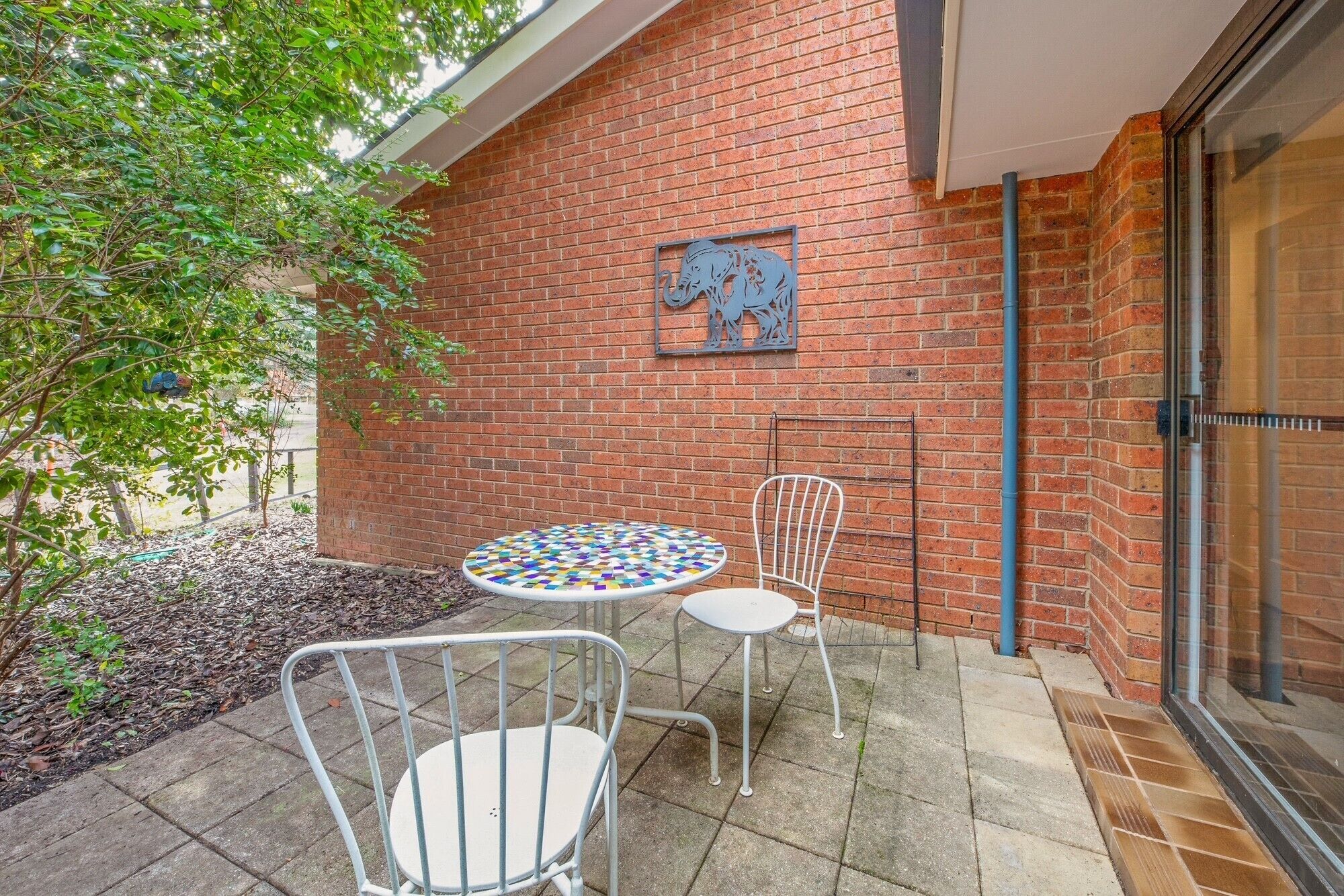 A patio area with a brick wall, a decorative metal elephant sculpture, two metal chairs, and a mosaic-tiled table.