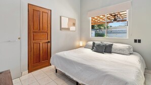 A bedroom with a wooden door, a bed with white bedding and gray pillows, a window with a view of greenery, and a ceiling fan. - 4BR Tropical Retreat • Pool • Beach • Golf Cart (Playa Potrero, Santa Cruz)