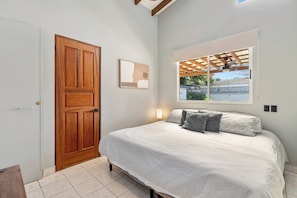 A bedroom with a wooden door, a bed with white bedding and gray pillows, a window with a view of greenery, and a ceiling fan. - 4BR Tropical Retreat • Pool • Beach • Golf Cart (Playa Potrero, Santa Cruz)