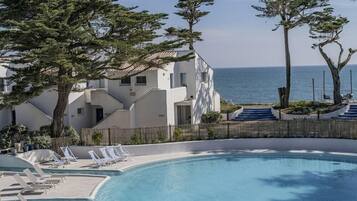 A pool area with lounge chairs, a clear view of the sea, and a building with a tiled roof.