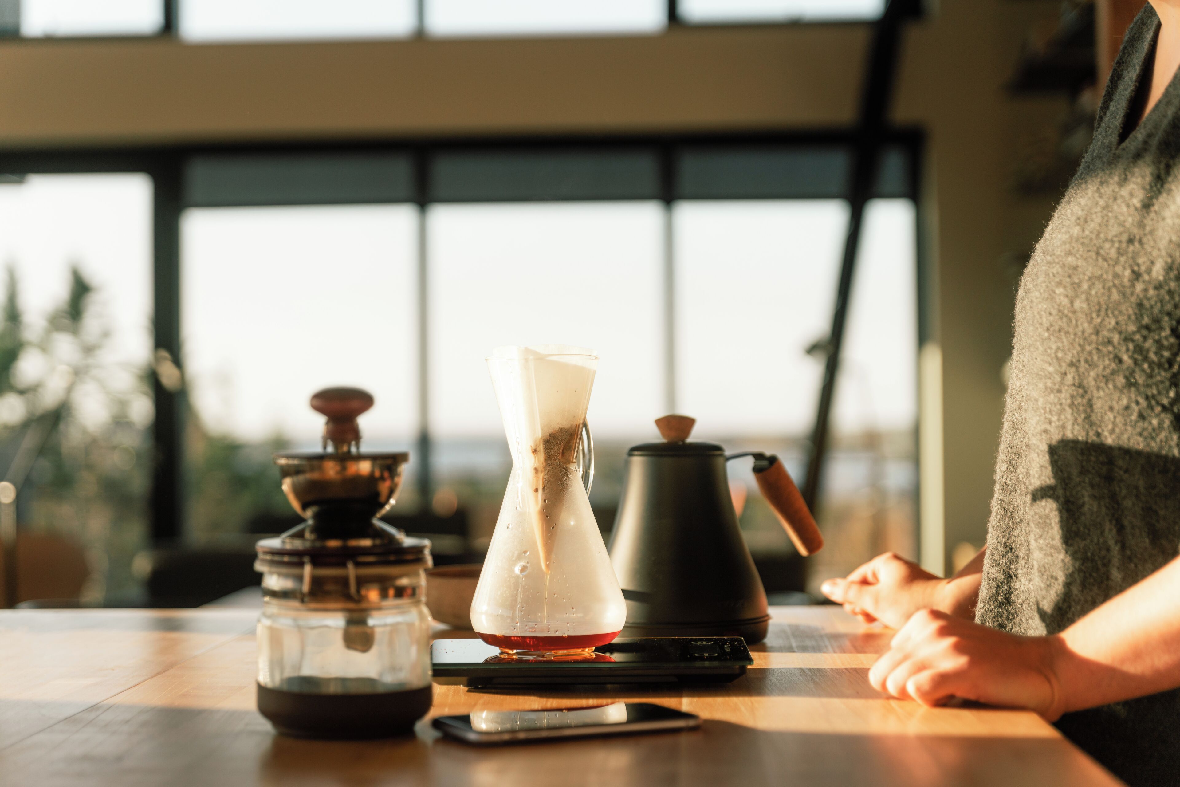 Una cafetera, una jarra de vidrio y un teléfono inteligente sobre una mesa de madera en una habitación con ventanas amplias.