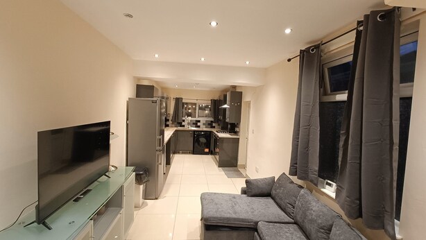 A modern kitchen with a built-in oven, a washing machine, and a dining area with a sofa and a TV. - Triplex Hot Tub House with Private Garden (Cardiff)