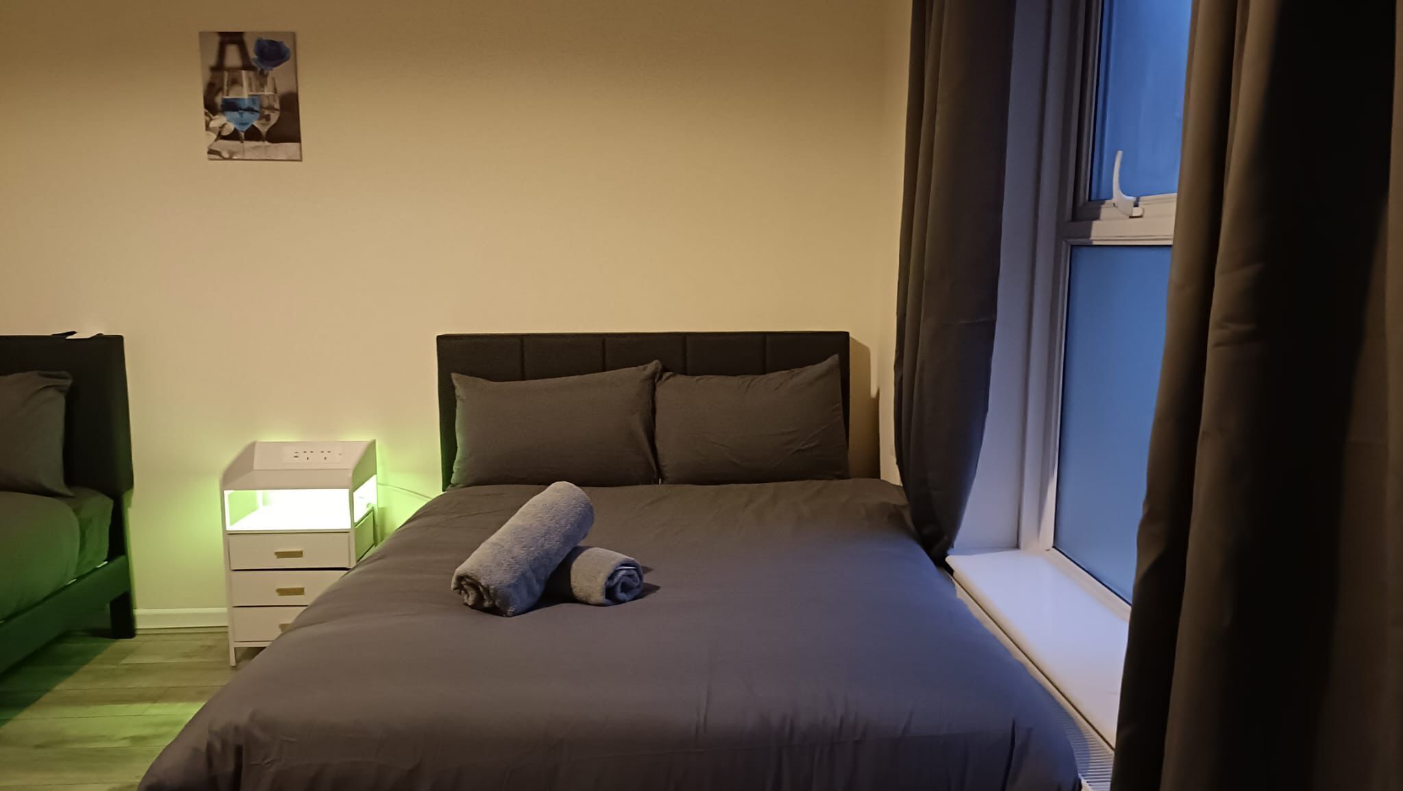 A neatly made bed with a grey blanket and two rolled towels, a bedside table with a lamp, a green chair, and a window with curtains.