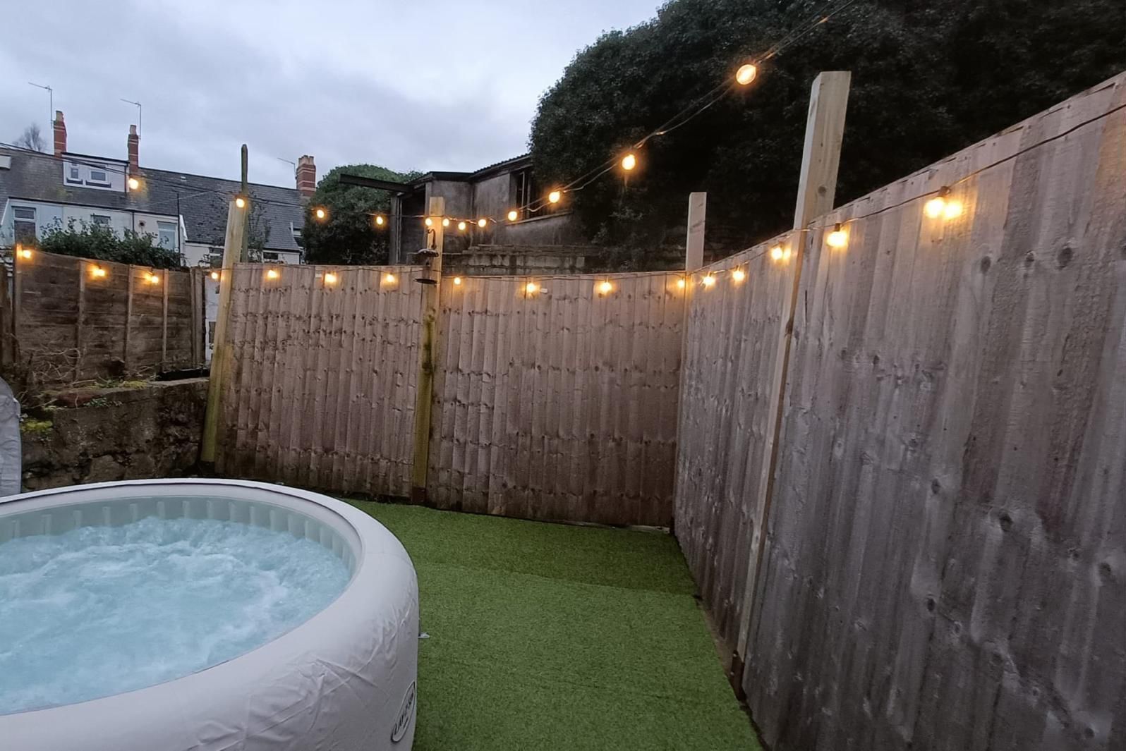 A backyard with a hot tub, artificial grass, wooden fence, and string lights.