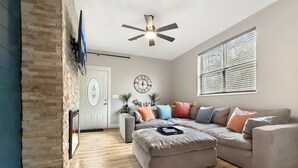 A modern living room with a stone accent wall, a ceiling fan, a large window with blinds, and a grey sectional sofa with multiple cushions. - Two Homes walk to Convention Ctr Hot Tub Game room (Branson)