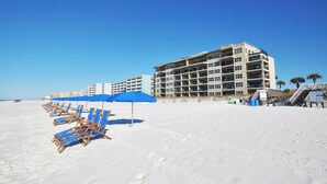 A beach with rows of blue umbrellas and lounge chairs, a multi-story hotel building, and palm trees in the background. - Emerald Towers West • Beach Front • Ocean View • Beach Service etw3003 (Fort Walton Beach)