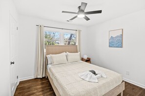 A bedroom with a bed, a ceiling fan, a window with curtains, a bedside table with a lamp, and a framed picture on the wall.