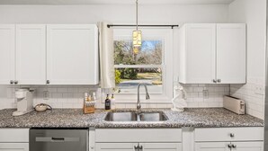 A modern kitchen with white cabinets, granite countertops, a built-in dishwasher, and a window with a view of a garden. - Spacious Charlotte Retreat with Private Hot Tub (Charlotte)