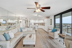A modern living room with a sofa, coffee table, and chairs, a kitchen in the background, and a balcony with a view. - Watercrest 1101 - Free Beach Chairs! (Panama City)
