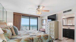 Un dormitorio con cama, ventilador de techo, televisión, cómoda y baño con espejo.