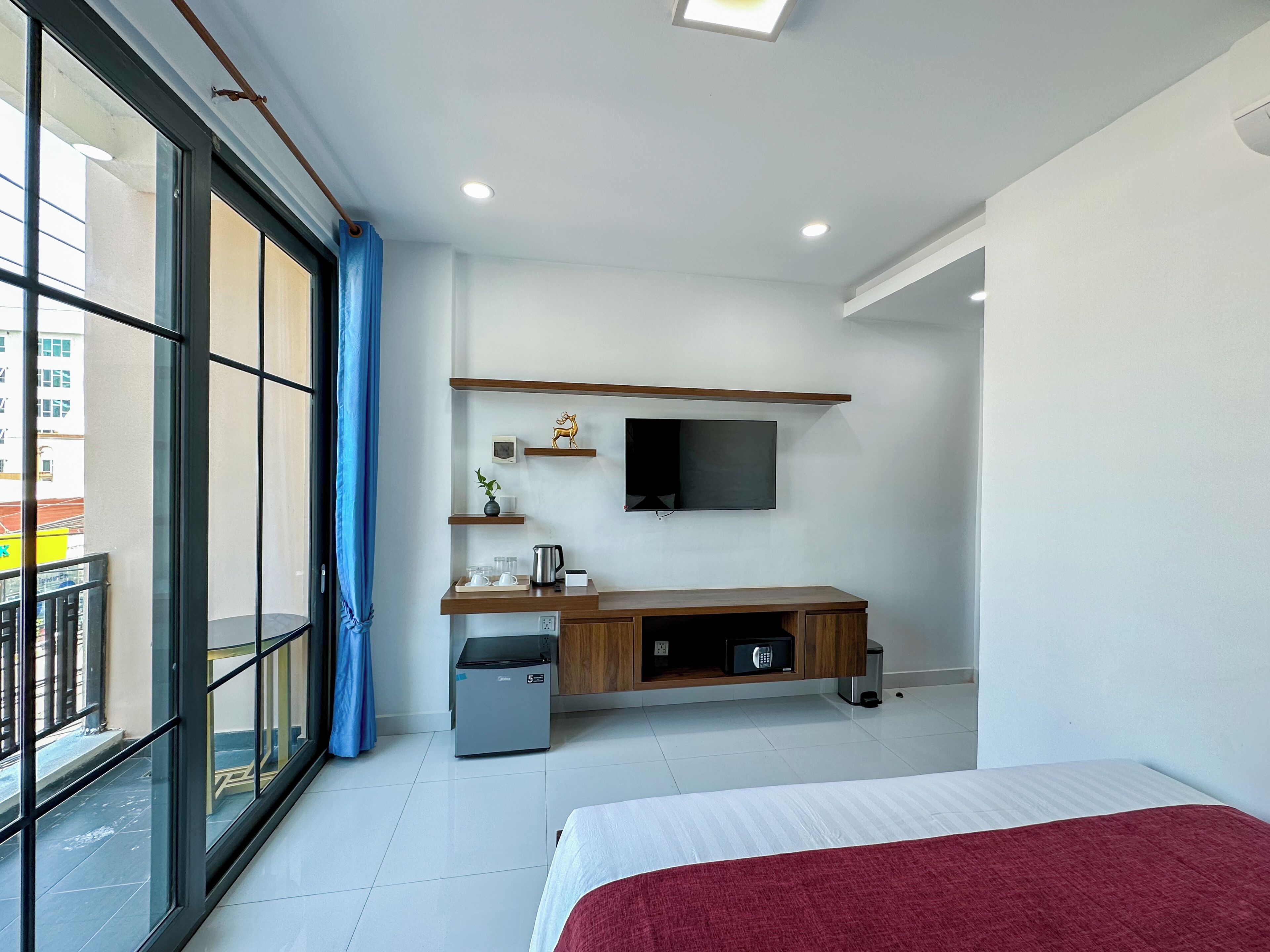 A hotel room with a bed, a desk with a flat-screen TV, a small kitchenette, and a window with a view of buildings.