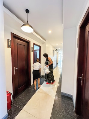 Een hotelgang met een brandblusser, een bagagekar en een persoon met een koffer.