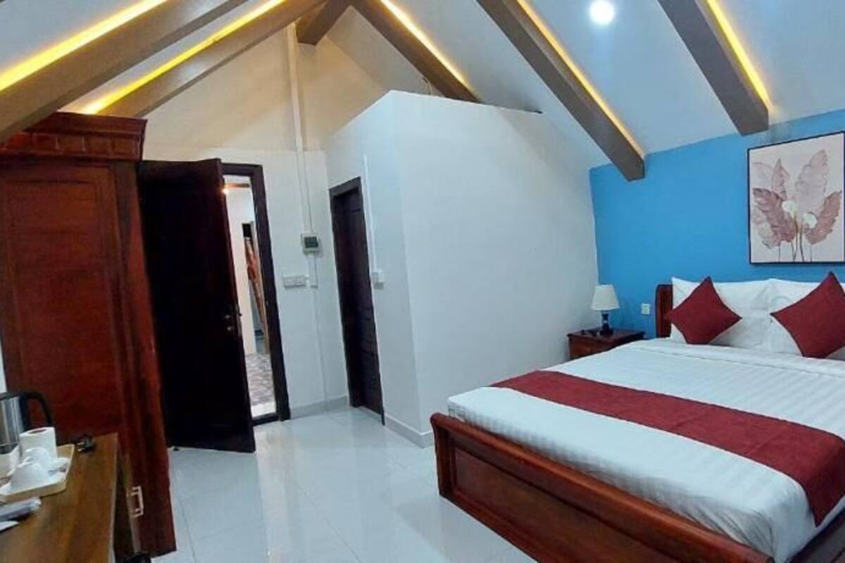 A bedroom with a large bed, wooden headboard, two bedside tables with lamps, a blue accent wall, and a ceiling with exposed beams.