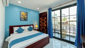 Quarto de hotel com uma cama grande, mesa de cabeceira, janela com vista da cidade e varanda com cadeiras.