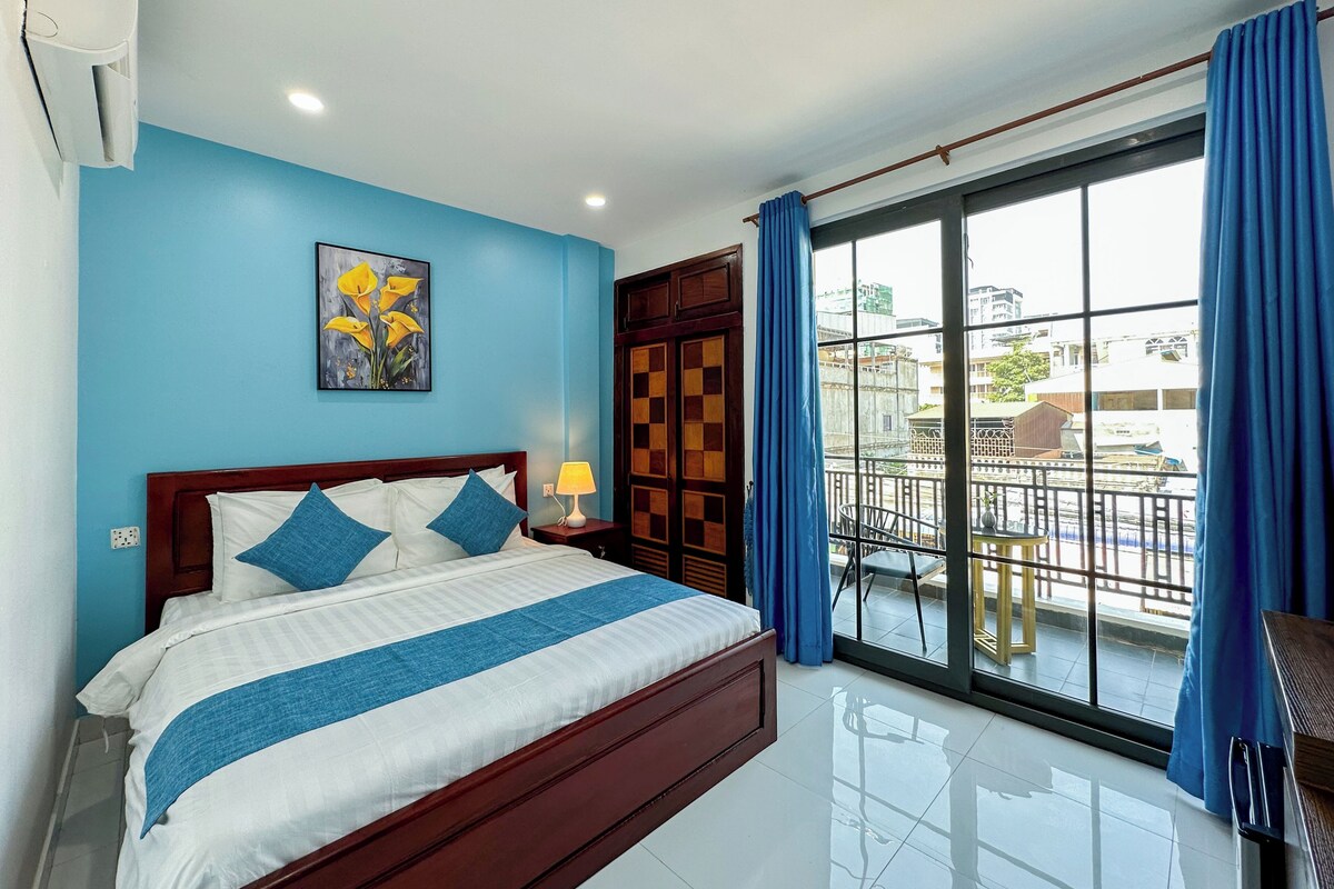 Habitación de hotel con una cama grande, una mesita de noche, un ventanal con vistas a la ciudad y un balcón con sillas.