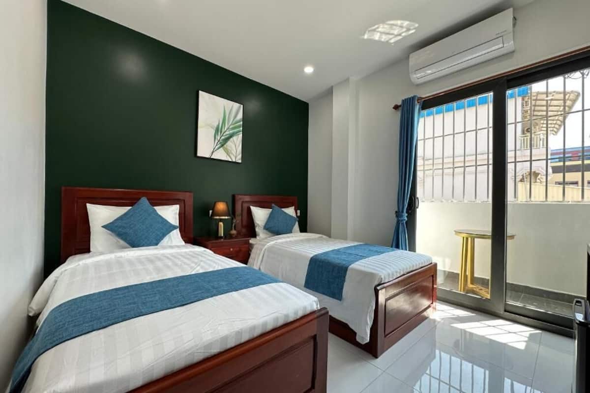A hotel room with two beds, a large window with curtains, a wall-mounted air conditioner, and a small table by the window.