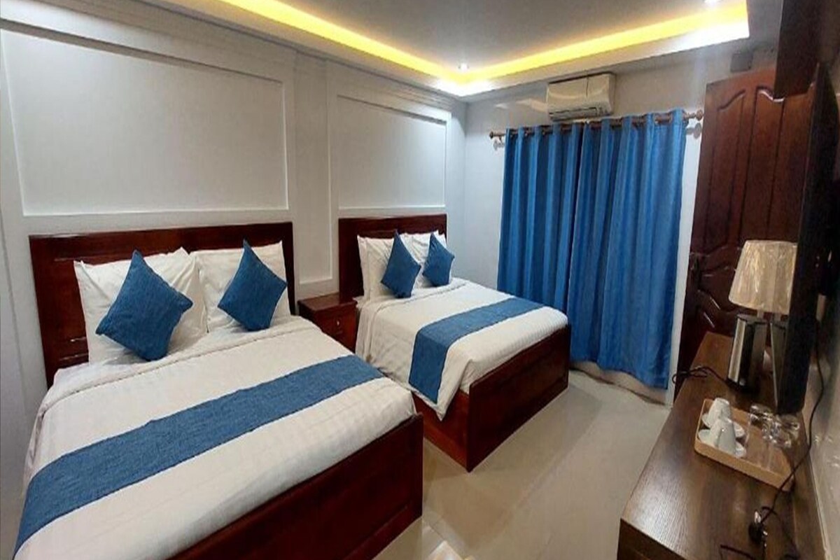 Habitación de hotel con dos camas, un escritorio con lámpara y una ventana con cortinas azules.