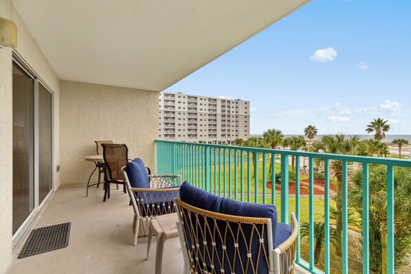 A balcony with wicker chairs and a table, overlooking a landscaped area with palm trees and a building in the background. - Gulf Front 2/2 Flash sale 3rd floor unit PPE6311 (Gulf Shores)