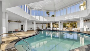 Uma piscina interior ampla, com vista direta para o céu através de grandes janelas, cercada por pilares brancos e um chão revestido de azulejos.