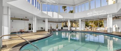Una amplia piscina interior con vista despejada al cielo a través de grandes ventanales, rodeada de pilares blancos y un suelo de baldosas.