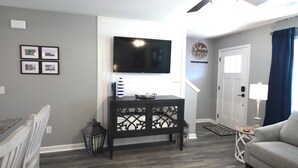 Ein Wohnzimmer mit einem Flachbildfernseher, einem dunklen Holzkonsolentisch, einem weißen Sessel, einem grauen Sofa und einem Deckenventilator.