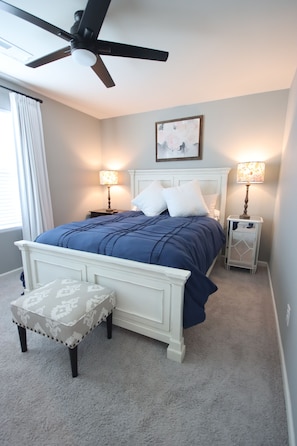 Une chambre à coucher comprenant un lit, deux tables de chevet, une commode, un ventilateur de plafond, une fenêtre avec des rideaux et un petit tabouret.
