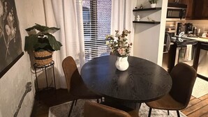 Ein Essbereich mit einem runden Tisch, Stühlen, einer Blumenvase und einer Pflanze auf einem Beistelltisch. Im Hintergrund ist eine Küche zu sehen.