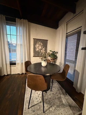 A dining area with a round table, four chairs, a vase with flowers, a painting on the wall, and a window with blinds. - Smokehouse 409 (Richmond)