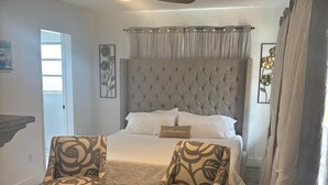 Quarto com cabeceira estofada, duas poltronas estampadas, cama com roupa de cama branca e ventoinha no teto.